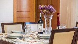 Ruokapöytä on katettu: siellä on lautasia, laseja, pullo Vina Belje Merlot -viiniä ja maljakko, jossa on kuivattuja kukkia.