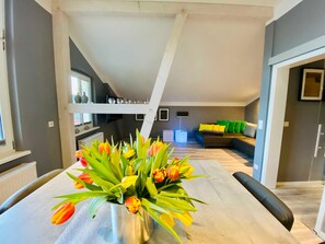 Un salon moderne avec une table à manger au premier plan, un canapé agrémenté de coussins colorés et un escalier menant à un niveau supérieur.
