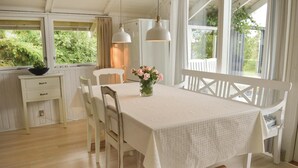 Un coin repas avec une table et un banc blancs, deux lampes suspendues, un vase avec des fleurs et une fenêtre avec des rideaux.