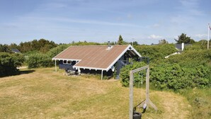 Une maison rustique, dotée d’un toit en pente, entourée d’un champ herbeux et d’une végétation dense.