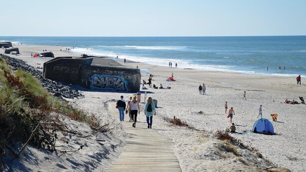 Une plage avec une promenade en bois menant au rivage, un grand rocher couvert de graffitis, et des gens qui se promènent et se détendent.