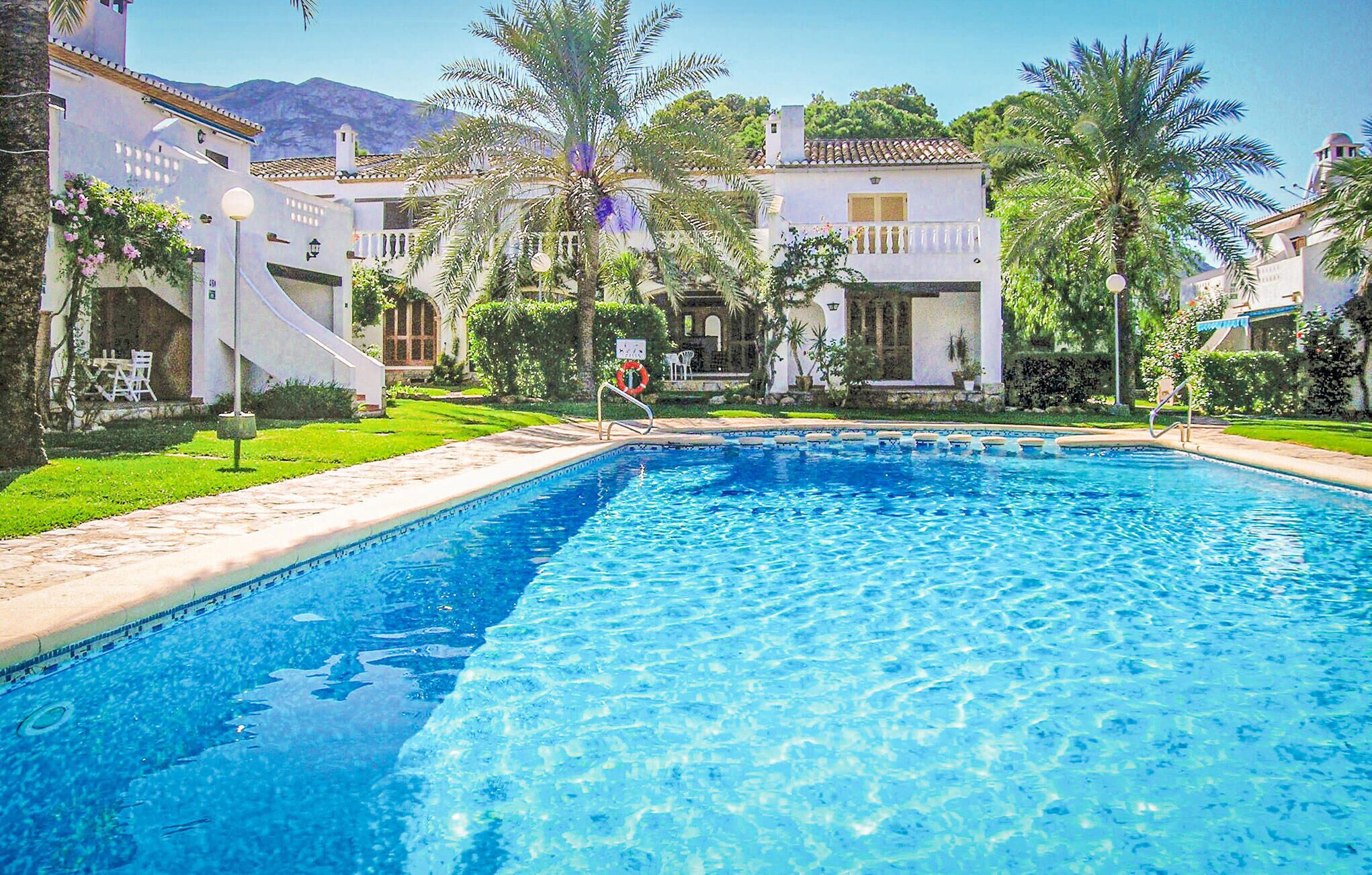 A swimming pool surrounded by white buildings with arched windows, palm trees, and a mountainous backdrop.