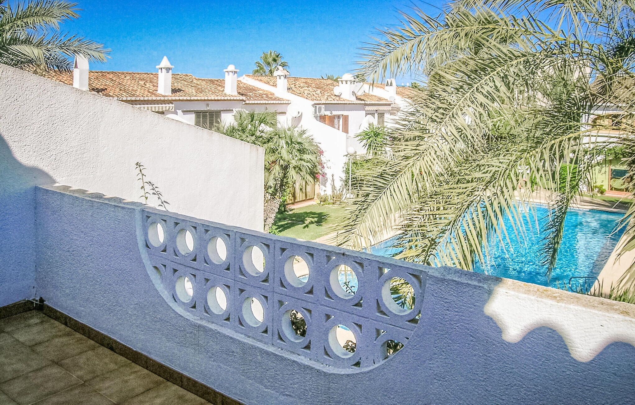 A poolside view with a clear blue pool, white buildings with red-tiled roofs, and palm trees.