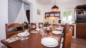 A dining area with a wooden table set for a meal, surrounded by wooden chairs. There is a kitchen area with modern appliances and a hanging light fixture above the table.