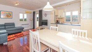 A modern living room with a sofa, a dining table with chairs, a TV, and a kitchen area with cabinets and a window.