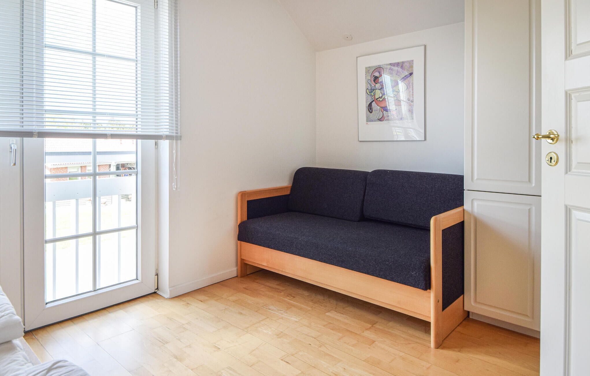 Une pièce avec un plancher en bois, un canapé gris foncé, une porte blanche avec une poignée dorée et une fenêtre avec des stores.