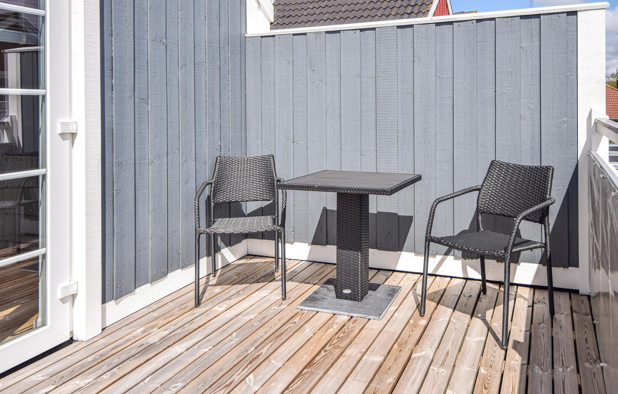 En träveranda med ett bord och två stolar, en grå vägg och ett fönster.