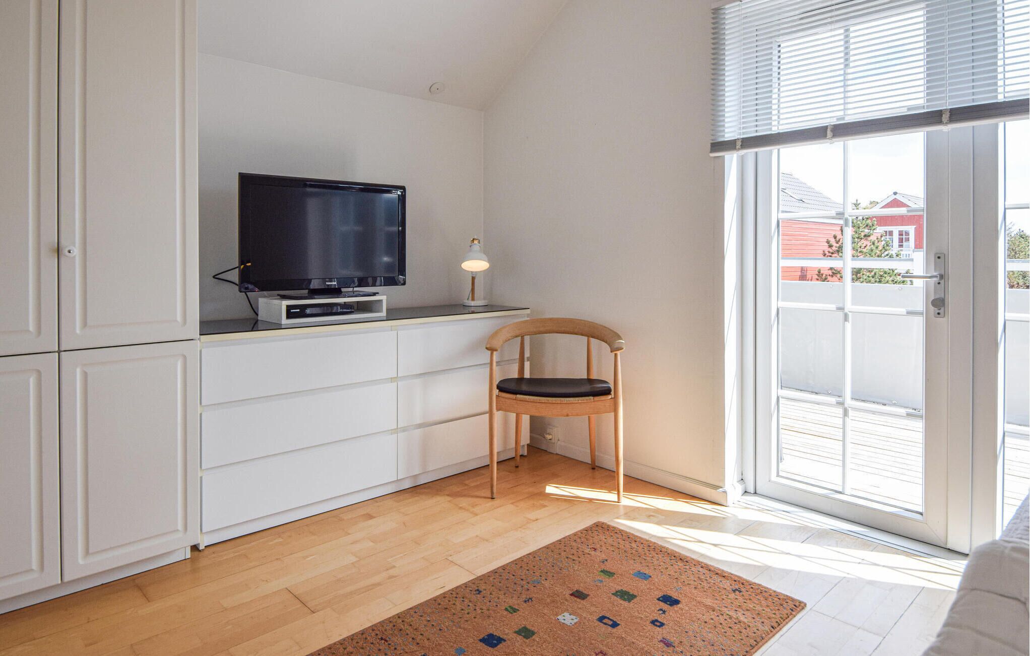 A room with a television on a cabinet, a wooden chair, a small table with a lamp, and a view through a sliding glass door.