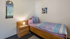 A bedroom with a wooden bed, a bedside table with a lamp, a small drawer unit, a window with blinds, and a wall-mounted picture. - Holiday apartment with shared balcony (Todtmoos)
