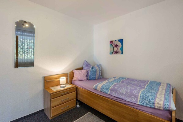 Une chambre avec un lit en bois, une table de chevet avec une lampe, une petite commode, une fenêtre avec des stores et un tableau accroché au mur.