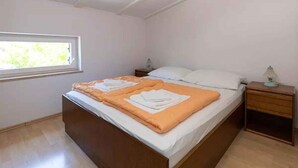 Una cama individual con una manta naranja, almohadas blancas, una mesita de noche de madera con una lámpara y una ventana con vistas a zonas verdes.