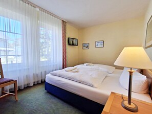 Une chambre d’hôtel avec un lit, une chaise, une table de chevet avec une lampe et une fenêtre avec des rideaux.