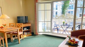A hotel room with a green carpet, a wooden desk with a TV, a bookshelf, and a dining table with fruit. - Vacation apartment 1.1.10 Inselstrand - Holiday apartment 1.1.10 Lottchen (Heringsdorf)