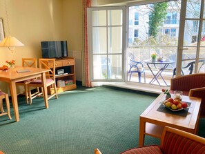 A hotel room with a green carpet, a wooden desk with a TV, a bookshelf, and a dining table with fruit.