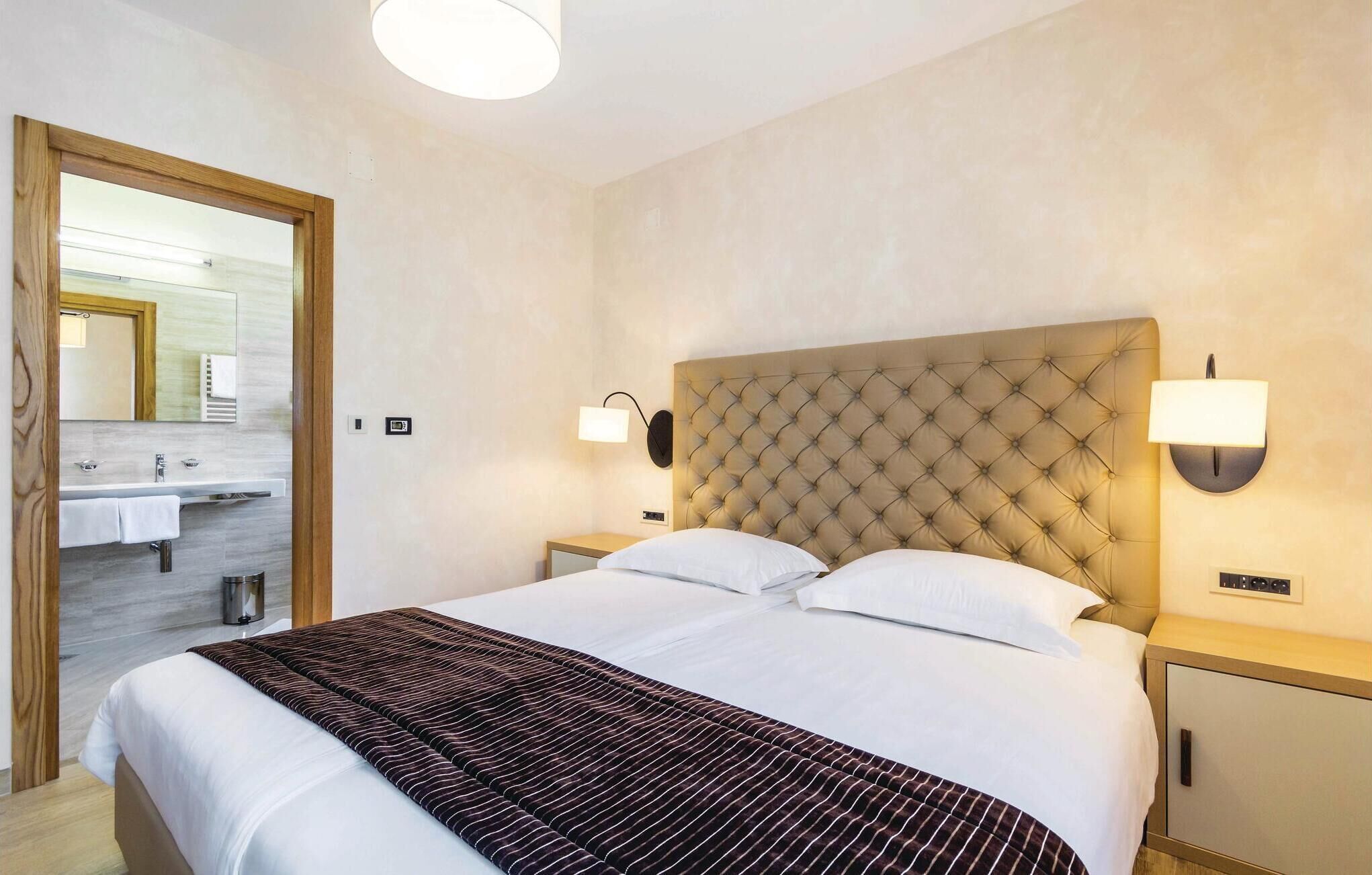 A hotel room with a large bed, two bedside tables, a headboard, and a bathroom visible through an open door.