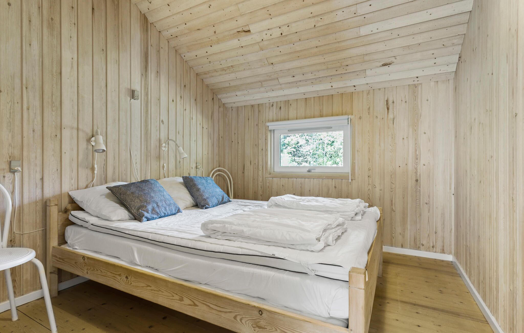 A single bed with white bedding and two patterned pillows in a room with wooden walls and a sloped ceiling.