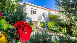 A two-story house with a red roof, surrounded by lush greenery and flowering plants. - Holiday apartment with a/c (Novi Vinodolski)