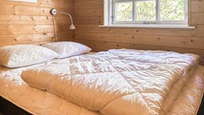 Sängyssä on pehmustettu patja ja tyynyt, yöpöydällä on lamppu ja ikkunasta avautuu näkymä puihin.