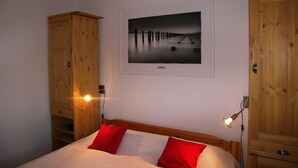 A bedroom with a wooden bed, two red pillows, a nightstand with a lamp, a wooden wardrobe, and a framed photograph on the wall. - Apartment 01 - Holiday home Knopf with 3 apartments, incl. WLAN (Zinnowitz)