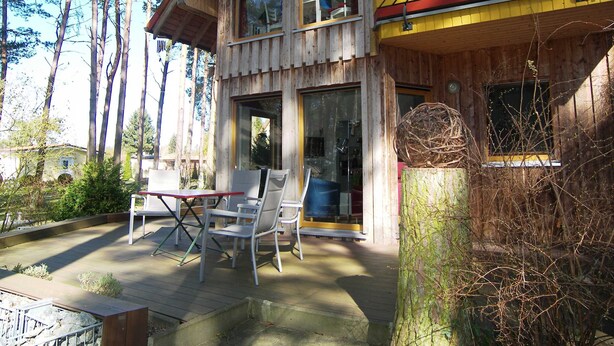 A wooden cabin with large windows, a deck with chairs, and a tree stump with a nest. - Apartment 01 - Holiday home Knopf with 3 apartments, incl. WLAN (Zinnowitz)