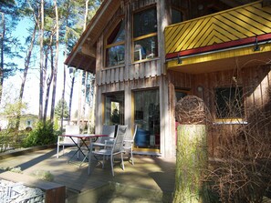 A wooden cabin with large windows, a deck with chairs, and a tree stump with a nest.