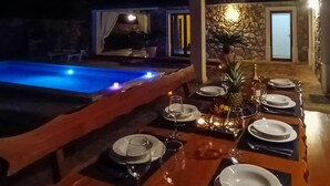A dining area with a poolside setting, including a table set for a meal, a stone wall, and a door leading to another room.