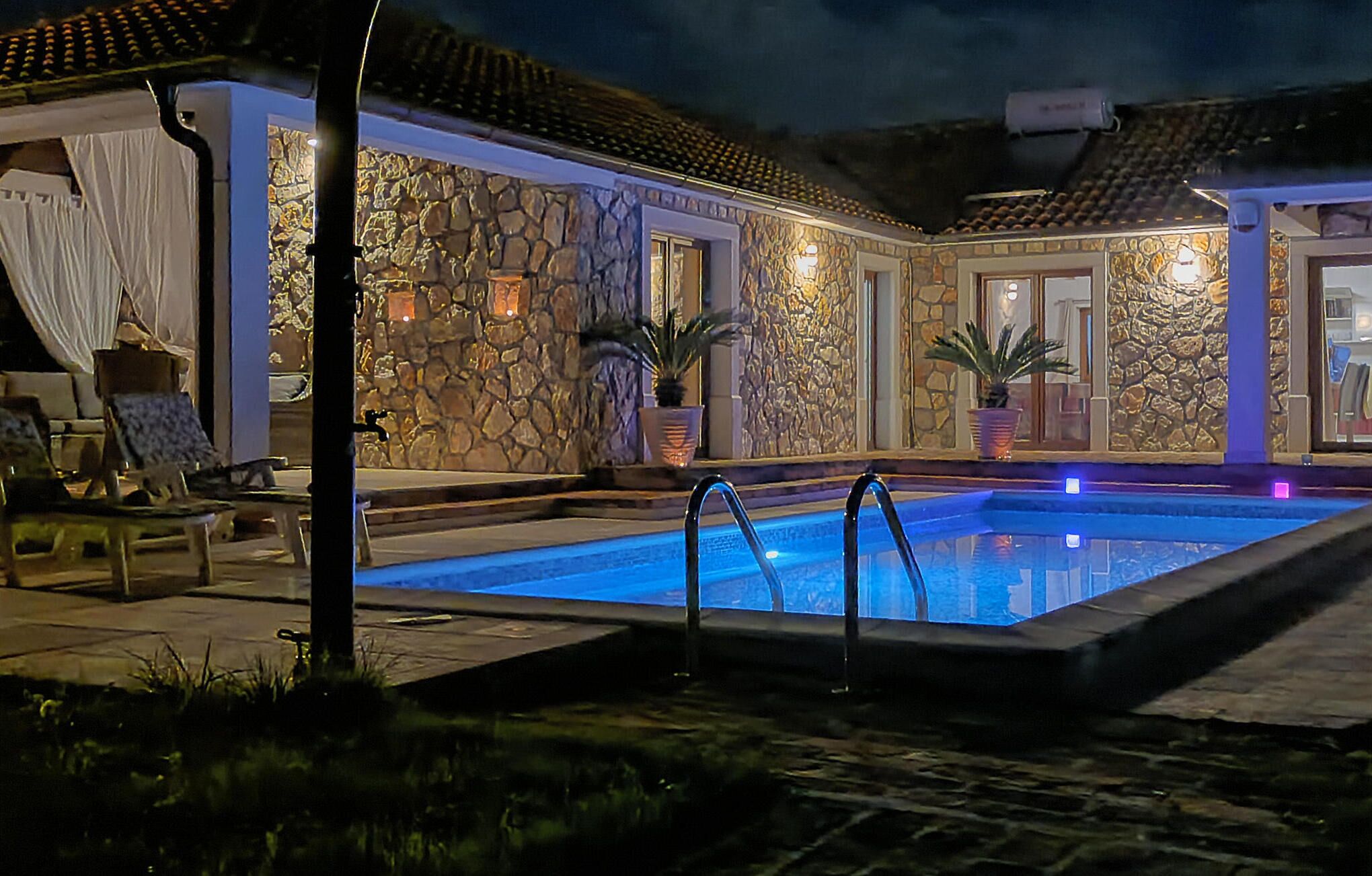 Una piscina con muro de piedra y techo de tejas, rodeada de sillas de descanso y plantas en macetas.