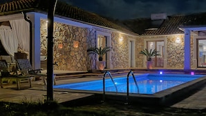 A swimming pool with a stone wall and tiled roof, surrounded by lounge chairs and potted plants.