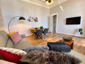 A modern living room with a sofa, a dining table with chairs, a TV mounted on the wall, and a hallway leading to another room.