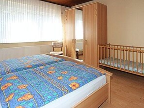 A bedroom with a bed, a wooden wardrobe, a crib, and a chair.