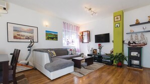 A modern living room with a sofa, coffee table, and a TV. There are paintings on the wall, a window with curtains, and a shelf with decorative items. - Amazing apartment in Bacinska jezera (Bacinska jezera)
