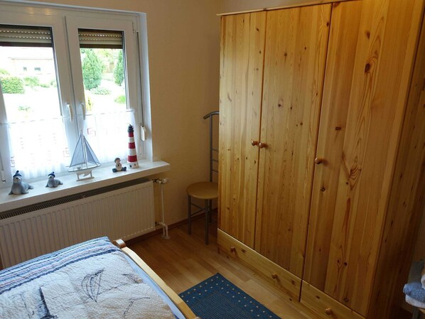 Et soveværelse med en stor træskab, et vindue med udsigt til grønne omgivelser, en radiator, en seng med ternet dynebetræk, og en lille stol.