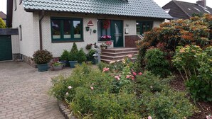 Une maison à deux étages, dotée d’un toit végétalisé, d’un porche à l’avant avec des marches et d’un jardin où poussent diverses plantes et fleurs.