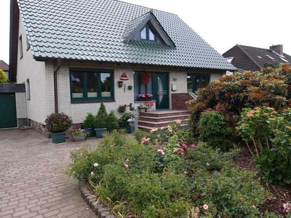 A two-story house with a green roof, a front porch with steps, and a garden with various plants and flowers. - Vacation apartment Heike, Germany (Rabel)