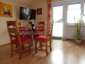 A dining area with a round table, four wooden chairs with red cushions, a bottle of wine, and two wine glasses. A television is mounted on the wall above the table.