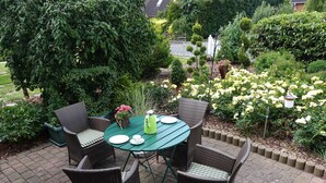 Une terrasse dotée d’une table ronde et de chaises en osier, entourée d’une végétation luxuriante et de plantes en fleurs.