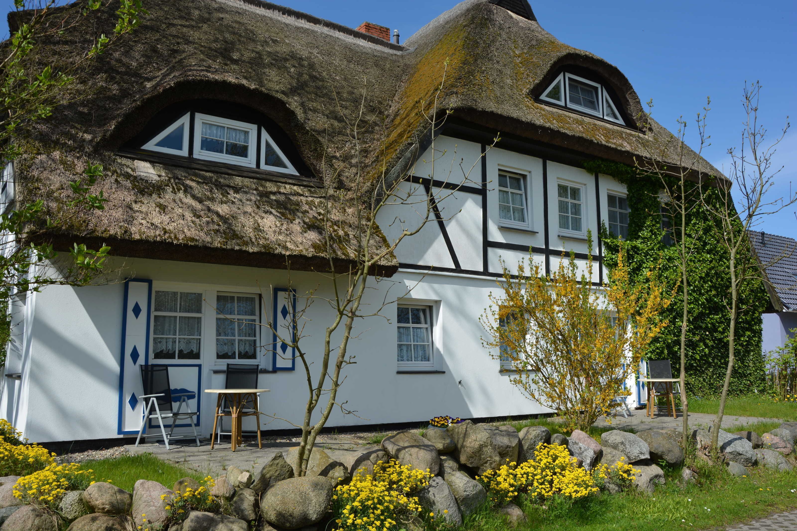 A traditional thatched-roof house with a white exterior, a small garden with yellow flowers, and a table and chairs outside.