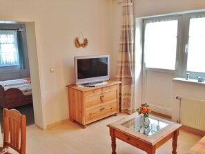 Une pièce comprenant un meuble TV en bois, une table basse avec un plateau en verre, un lit recouvert d’une couvre-lit à motifs blancs et rouges, et une fenêtre avec des rideaux.