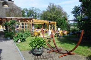 Un espace extérieur aménagé avec des tables et des chaises, entouré de plantes en pot et bordé d’un bâtiment rustique doté d’un toit de chaume.
