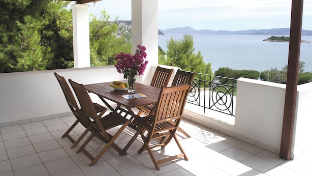 Un espace extérieur abrité, aménagé pour prendre les repas, avec une table et des chaises, un vase de fleurs et une vue sur la mer et les montagnes.