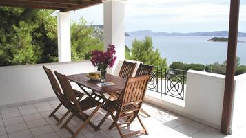 Ein überdachter Außenbereich zum Essen mit Tisch und Stühlen, einer Blumenvase und Blick auf das Meer und die Berge.