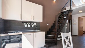 A modern kitchen with white cabinets, a stainless steel sink, and a built-in oven. A wooden staircase with a metal railing leads to a landing.