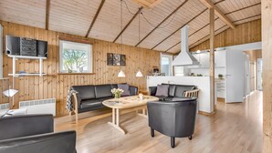 Ampla sala de estar com interior em madeira, um sofá de couro preto, uma mesa de centro redonda e uma cozinha ao fundo.