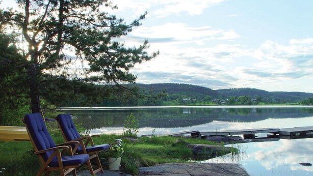 Two blue chairs by a calm lake with a dock, surrounded by greenery and a distant view of houses.