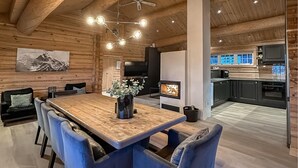 A modern log cabin interior with a wooden dining table, blue upholstered chairs, a fireplace, and a kitchen with dark cabinets and a large window.