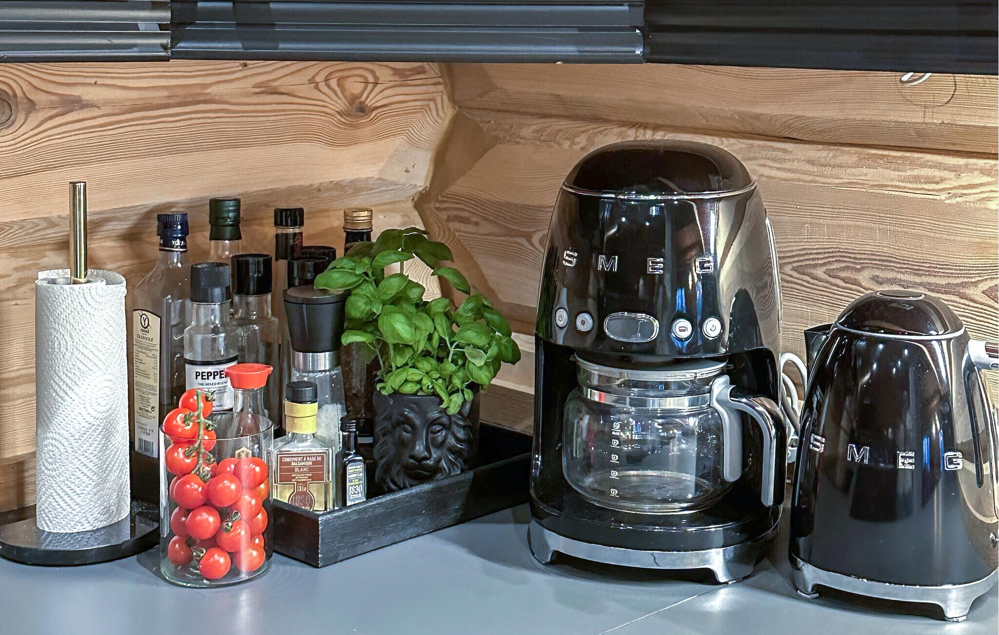 Een keukentafel met een koffiezetapparaat, een waterkoker, een glazen pot met tomaten en verschillende kruiden.