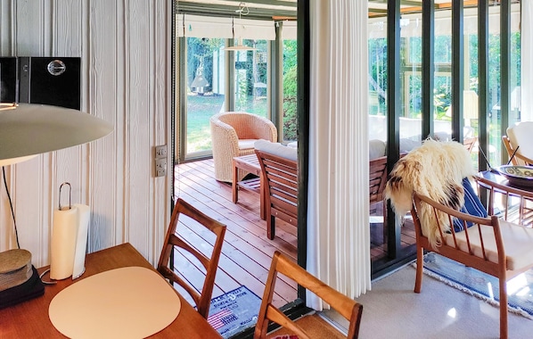 A room with a wooden table, chairs, a wicker sofa, and a view of the outdoors through large windows.