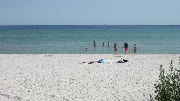 Une plage de sable blanc aux eaux calmes. On peut voir des gens dans l’eau et sur la plage.