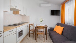A compact kitchen with white cabinets, a stove, and a sink. There's a dining table with chairs and a gray sofa with orange cushions. A flat-screen TV is mounted on the wall. - Gorgeous apartment in Porec with swimming pool (Porec)