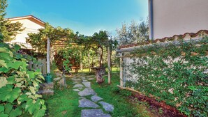 A stone pathway leading through a lush garden area with a pergola covered in vines, a rustic wooden bench, and a small stone table.